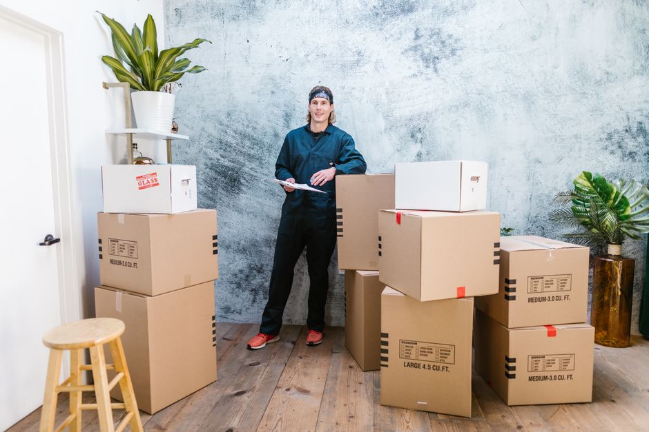 Smiling mover in coveralls standing with parcels, clipboard in hand, indoors.